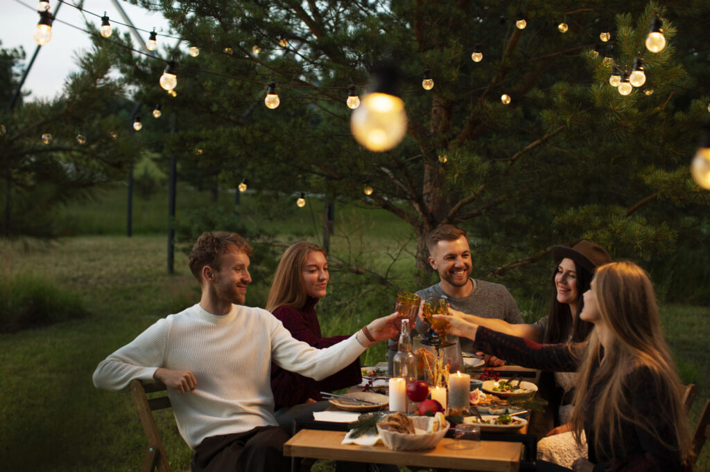Group of friends toasting at an outdoor garden party under warm string lights, with a festive table setting and trees in the background.