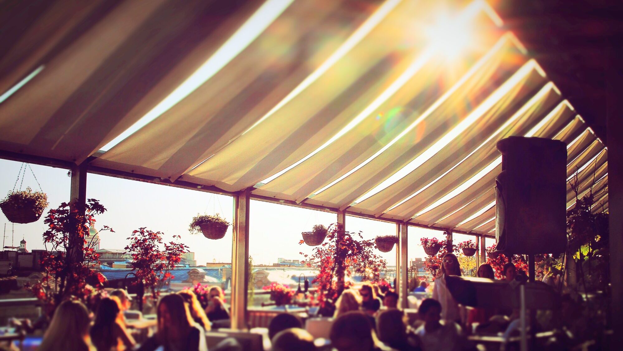 Daytime rooftop event with guests seated under a fabric-covered shelter, surrounded by hanging baskets and sunlit views of the city skyline.