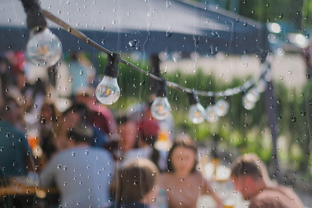 Raindrops on glass with blurred outdoor event and string lights in the background, capturing the challenge of unpredictable weather at UK events