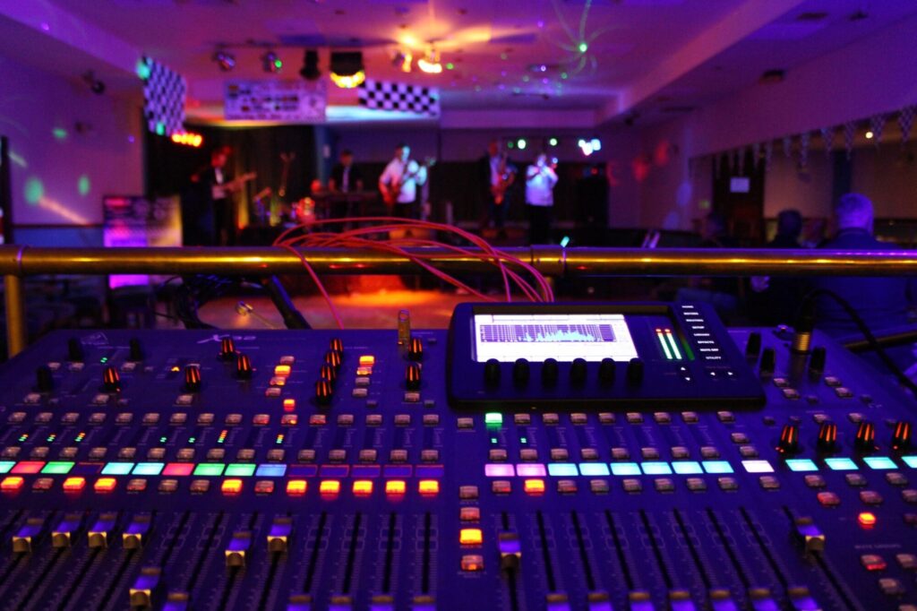Digital mixing desk glowing with colourful LEDs at a live indoor gig, with a band performing in the background—showcasing the sound control needed for music-focused events.
