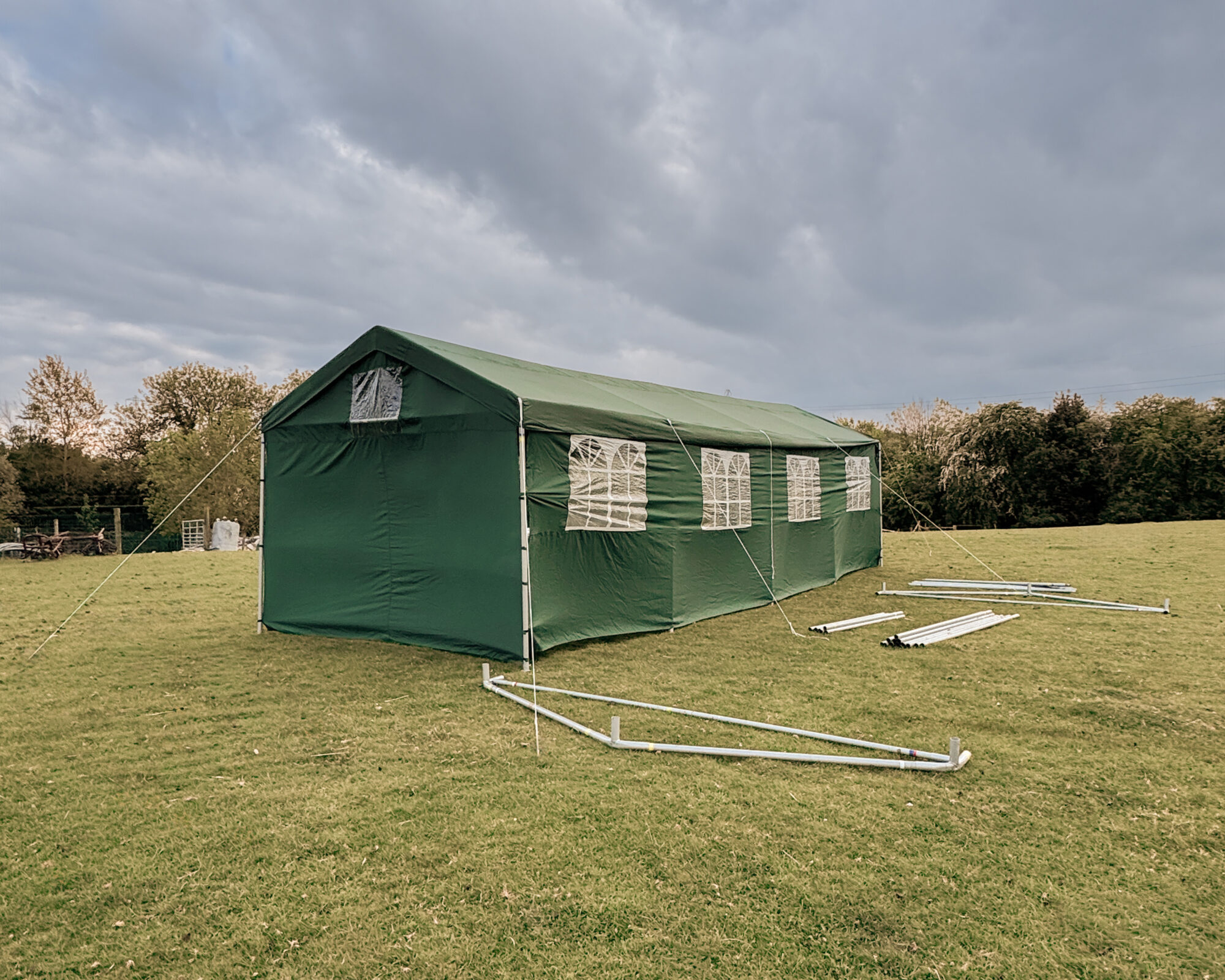 9m x 3m green event tent with side panels set up on a grass field under cloudy skies.