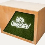 Rustic DJ booth with Let’s Celebrate neon sign and green grass-style front panel
