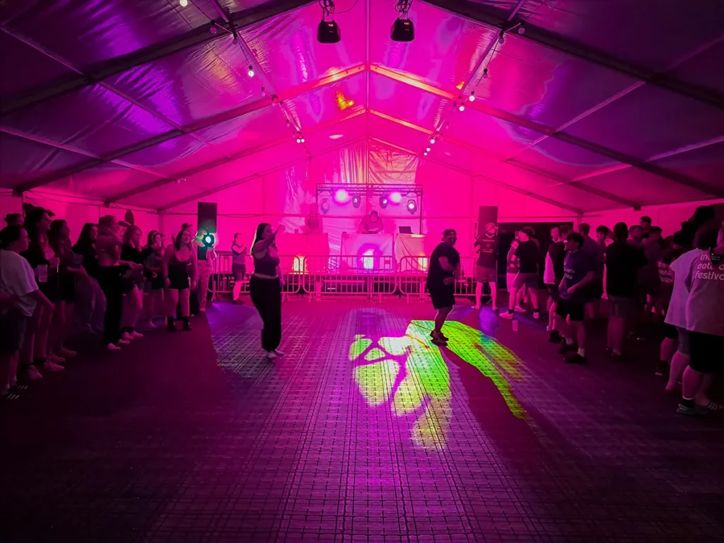 Silent disco dancefloor inside a marquee with pink lighting, DJ booth, stage lights and guests gathered around the floor.