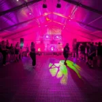 Silent disco dancefloor inside a marquee with pink lighting, DJ booth, stage lights and guests gathered around the floor.