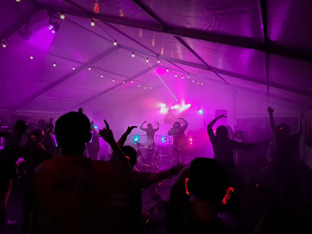 Silent disco crowd dancing under purple lighting inside a marquee with DJ booth, smoke effects and party lights.