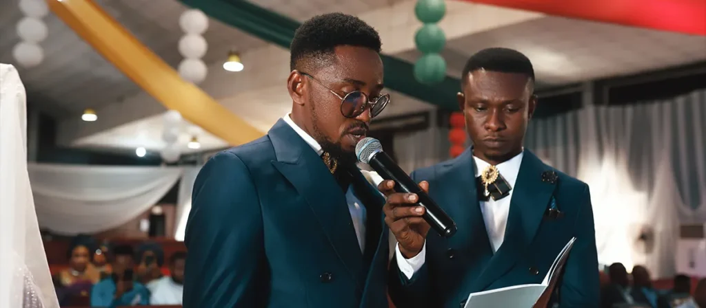 Groomsman reading a wedding speech into a handheld microphone during an indoor ceremony.