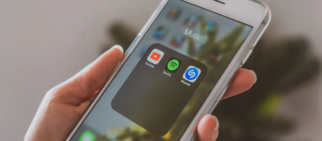 Hand holding a smartphone showing music apps including YouTube, Spotify and Shazam.