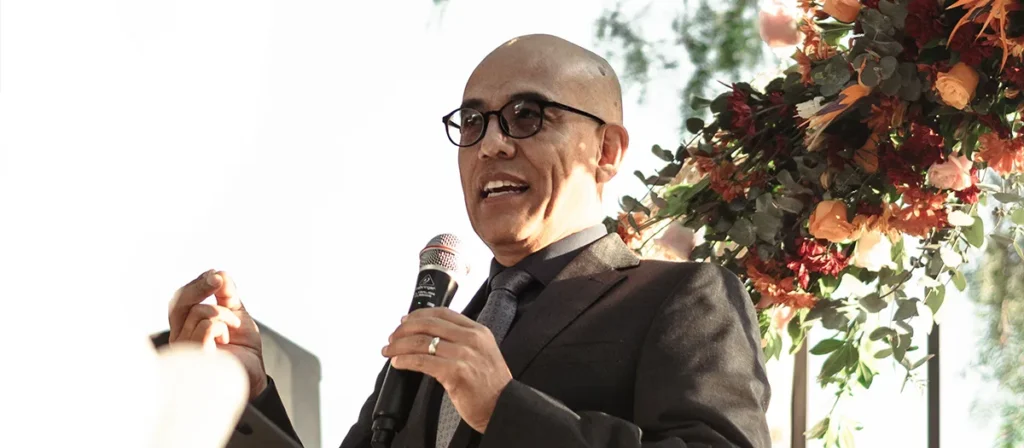 Man giving a speech with a handheld microphone at an outdoor wedding ceremony with floral decorations.