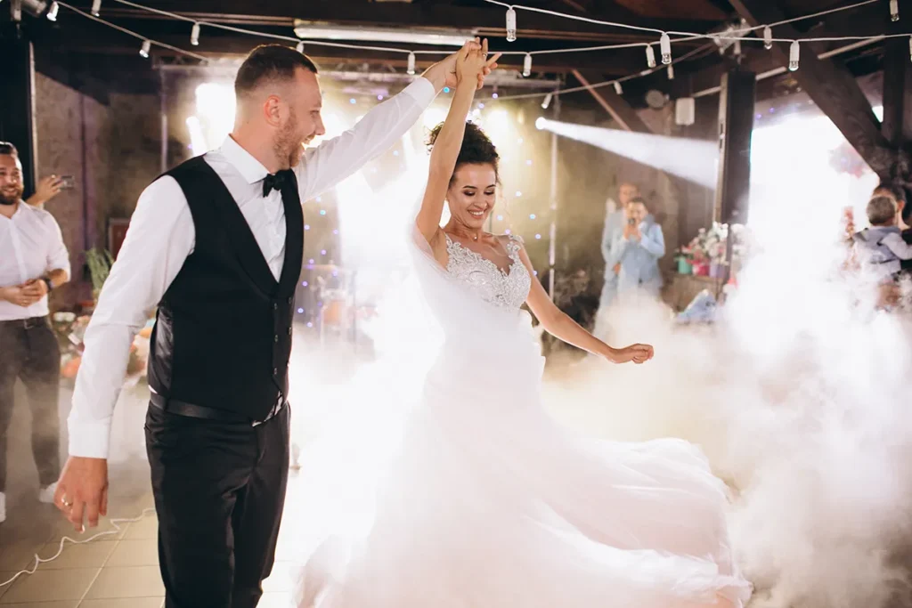 Bride and groom dancing on a smoky wedding dancefloor with dramatic lighting and guests in the background.